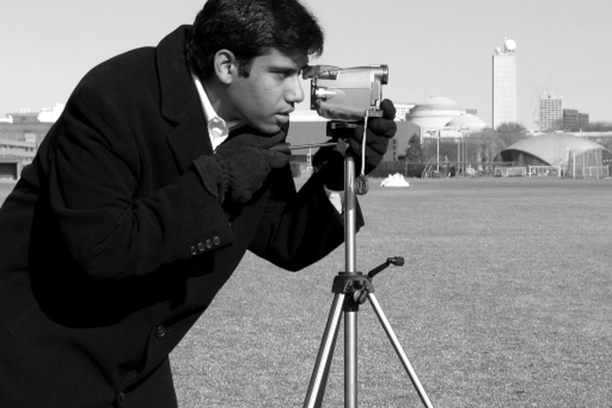 A photographer looking through a camera mounted on a tripod outdoors.